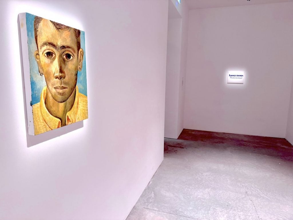 A painting of a man's face in a hall next to a small painting that says "ANONYMOUS HOMOSEXUAL"