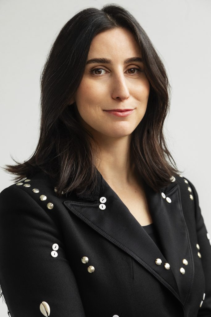 a headshot of a woman with medium length dark brown hair and brown eyes, wearing a black blazer with studs here and there
