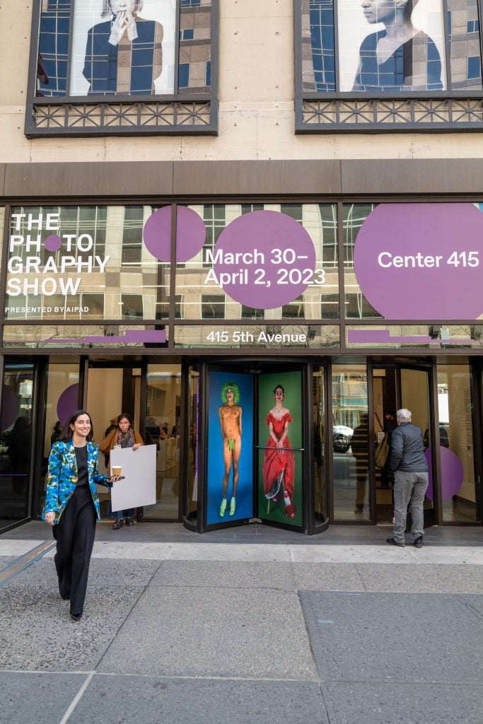 a woman in a blue blazer holding a paper cup of coffee and her cell phone and standing on the sidewalk in front of a building with signs for "the photography show presented by AIPAD" — on the revolving doors are two photographs on of a nude person in a green wig with a fig leaf covering their body and another of a woman in a red dress that looks like its from another century