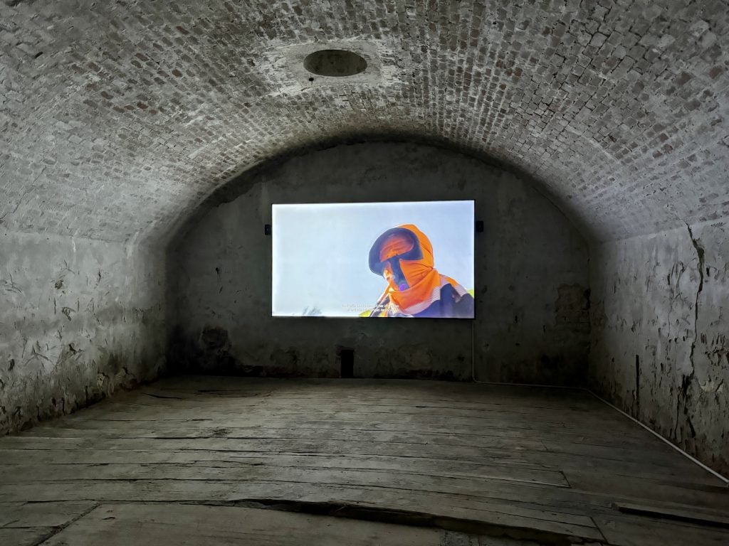 A film of a person in an orange hood plays within a stone chamber