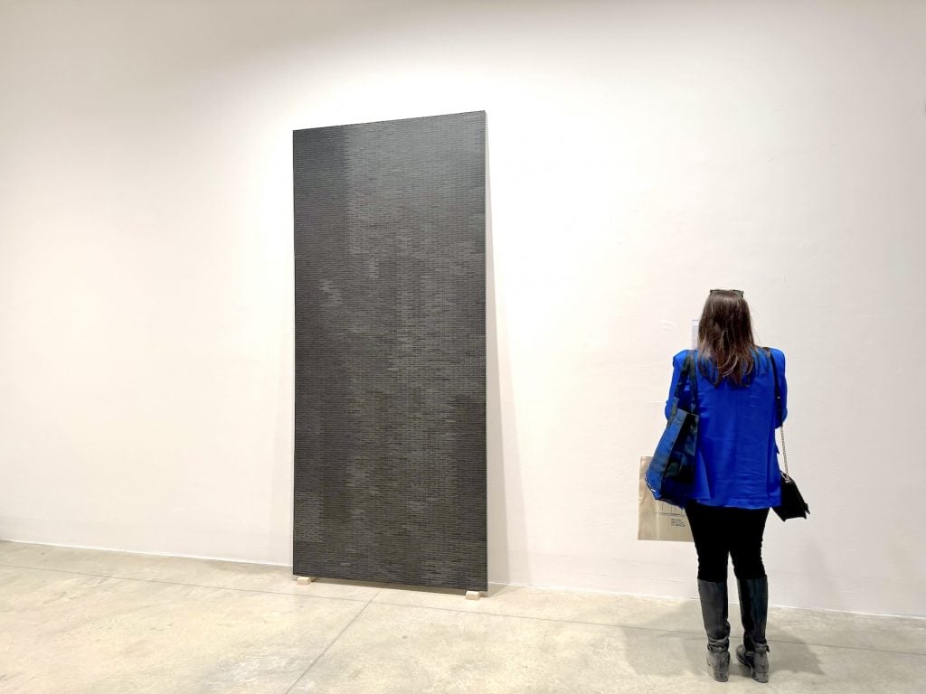 A woman looks at a wall label next to a densely gridded black abstract painting leaned against the wall