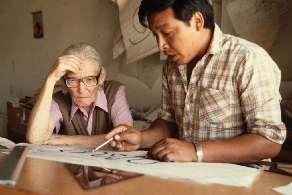 an old woman wearing glasses leans over a table, watching a man to her right drawing at a desk