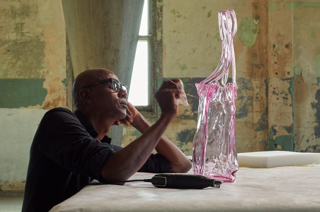 The artist Mark Bradford leans against a table etching on a pink glass sculpture.