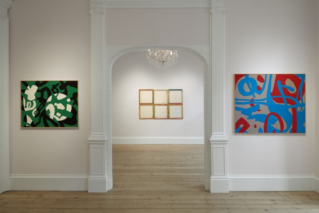Installation view of an exhibition with an abstract painting on either side of a traditional molded doorway through which another artwork is hung on a back wall, and a glimpse of a crystal chandelier overhead.