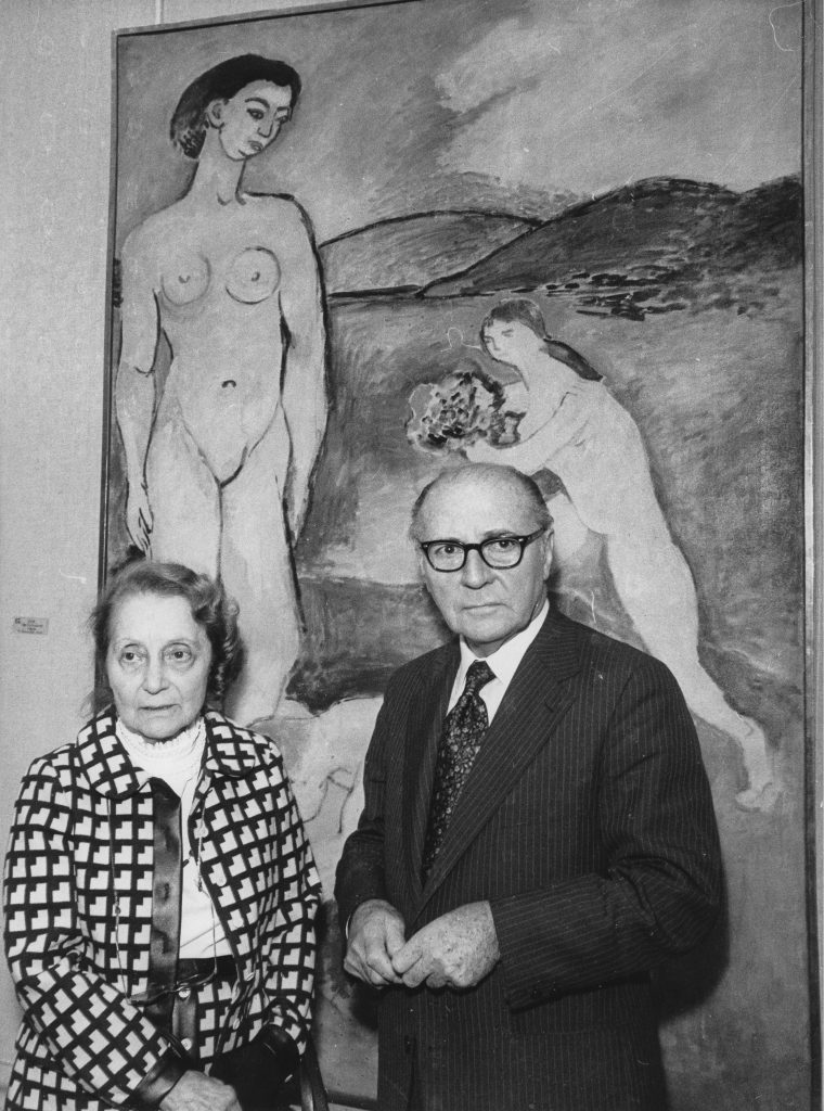 A short woman stands on the left and a man in a suit to the right. They are standing in front of a painting of two nude women by Matisse