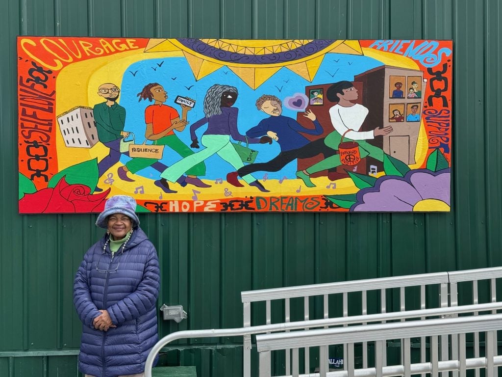 A photo of a Black woman in a blue coat and hat standing and smiling before a colorful mural installed on a deep green wall
