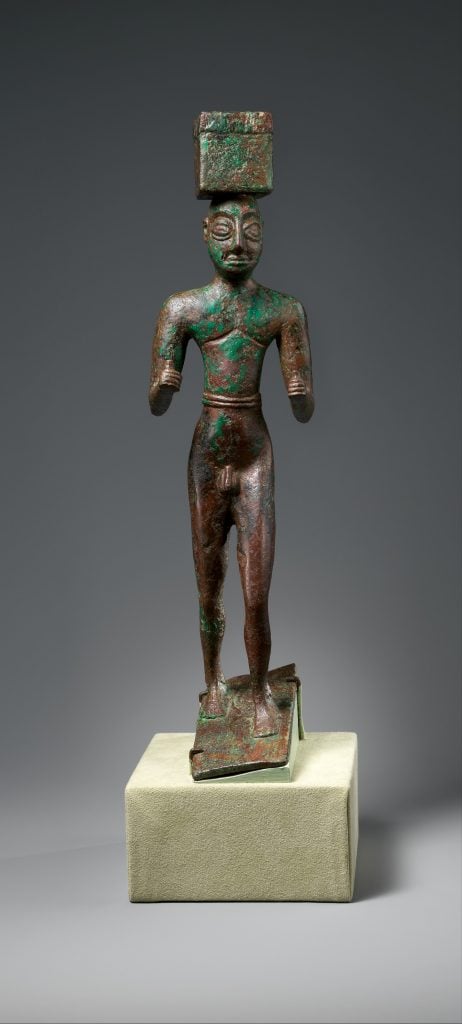 A bronze sculpture from antiquity, mounted on a museum display block. The figure stands upright, with pronounced muscles, and wears a high, squared headdress. It exhibits a green patina indicative of aged copper alloys, such as bronze. The stance is rigid, with fists clenched by the sides and legs together. This artifact conveys a sense of historical significance and cultural heritage, likely from the Middle Eastern region. The grey background emphasizes the statue's texture and color, highlighting its age and the fine craftsmanship of a bygone era.