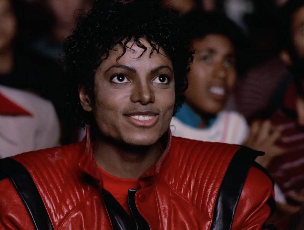 A man, pop star Michael Jackson, sitting in a darkened theater wearing a red jacket.