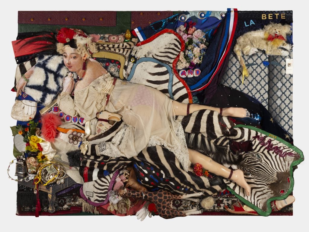 An overhead look at a woman, made of doll legs and felt, laying on her stomach looking back up at the viewer with mixed media surrounding of objects such as purse, gloves, fake flowers, hat, toys, and stuffed animals.