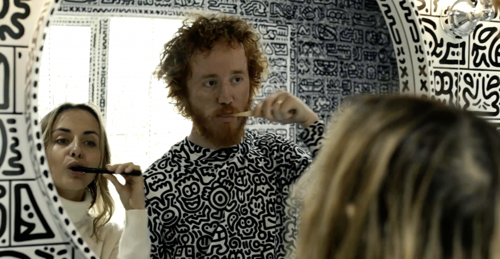 A man and a woman brush their teeth in a bathroom covered in doodled drawings