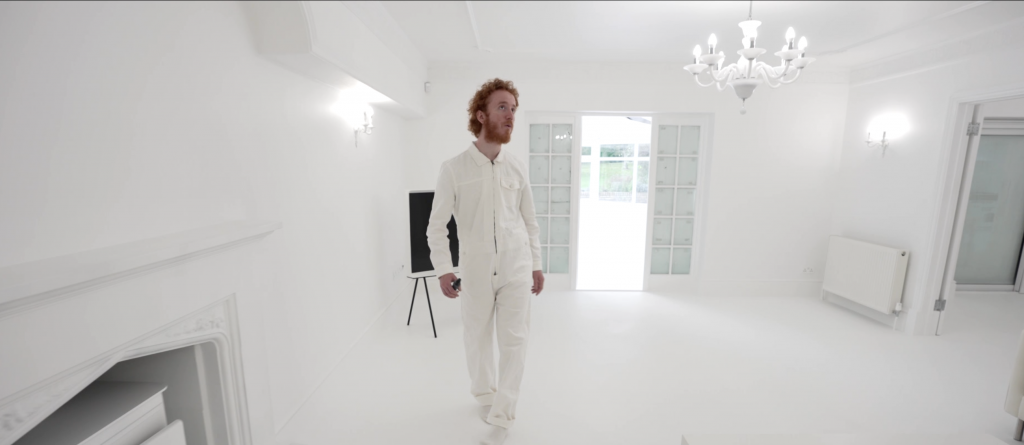 An artist in a white jumpsuit stands in a vacant white room