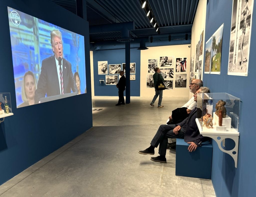 In an art gallery, two people watch a video of Donald Trump speaking