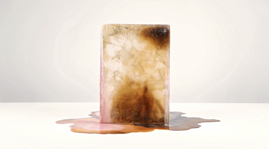 A photo of an amber-tinged ice block melting on a white table before a white background