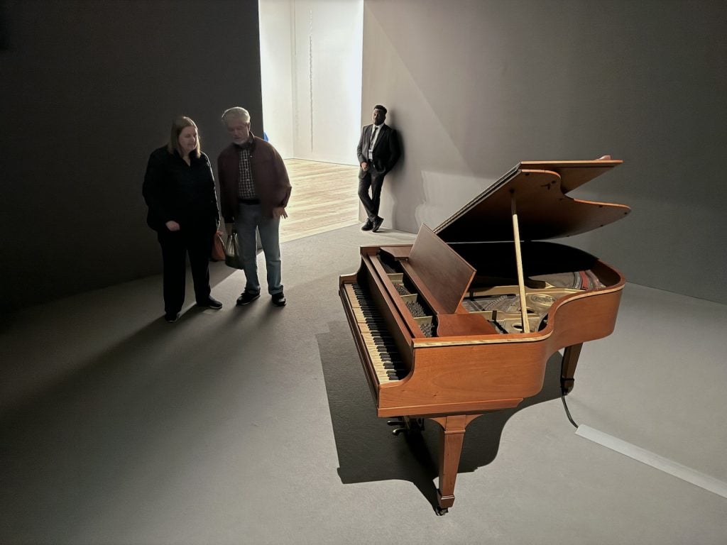 Two visitors and a guard look at a piano in a darkened art gallery