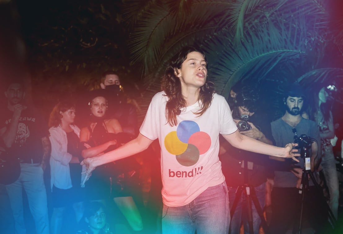 Artist Nora Turato wearing jeans and a graphic t-shirt in front of a crowd of people speaking, the image has light flares of blue, green, and hot pink.