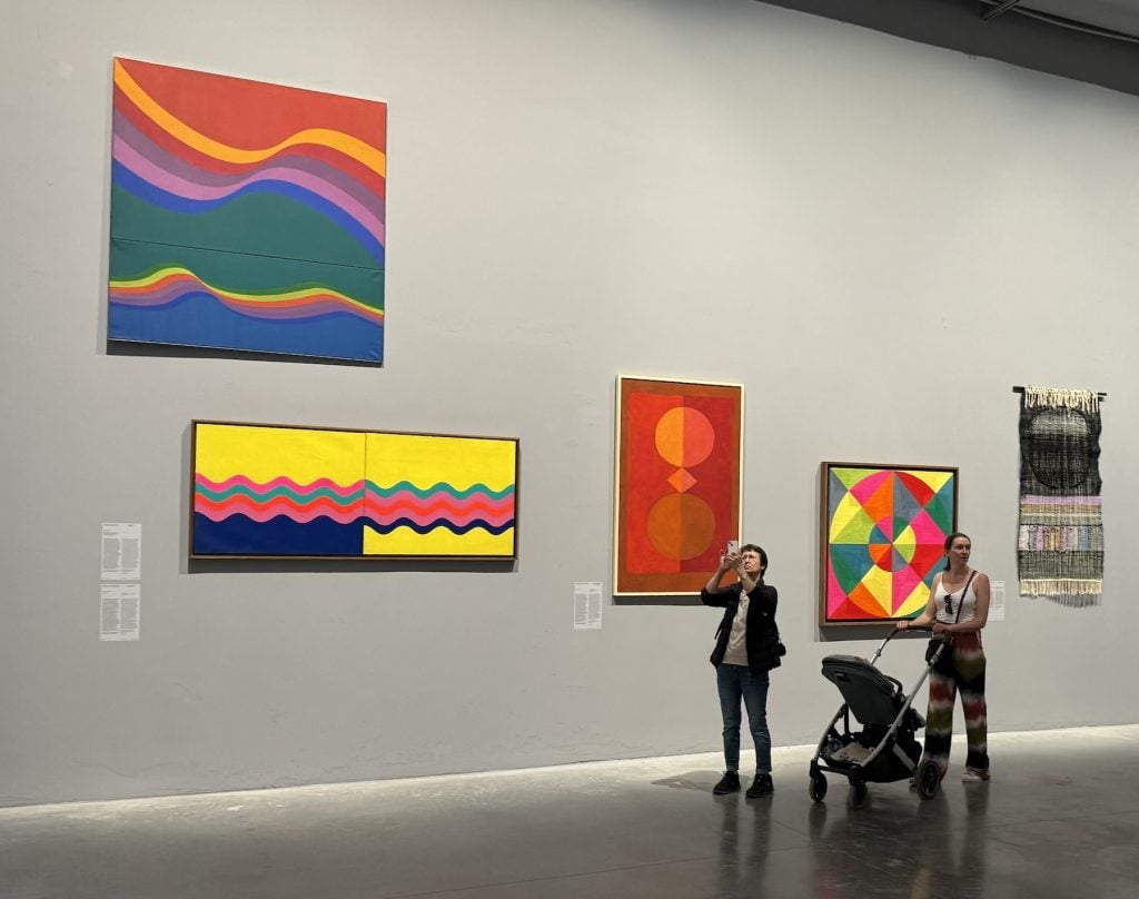 A woman takes a photo and a woman pushes a stroller in front of a wall of colorful abstract paintings