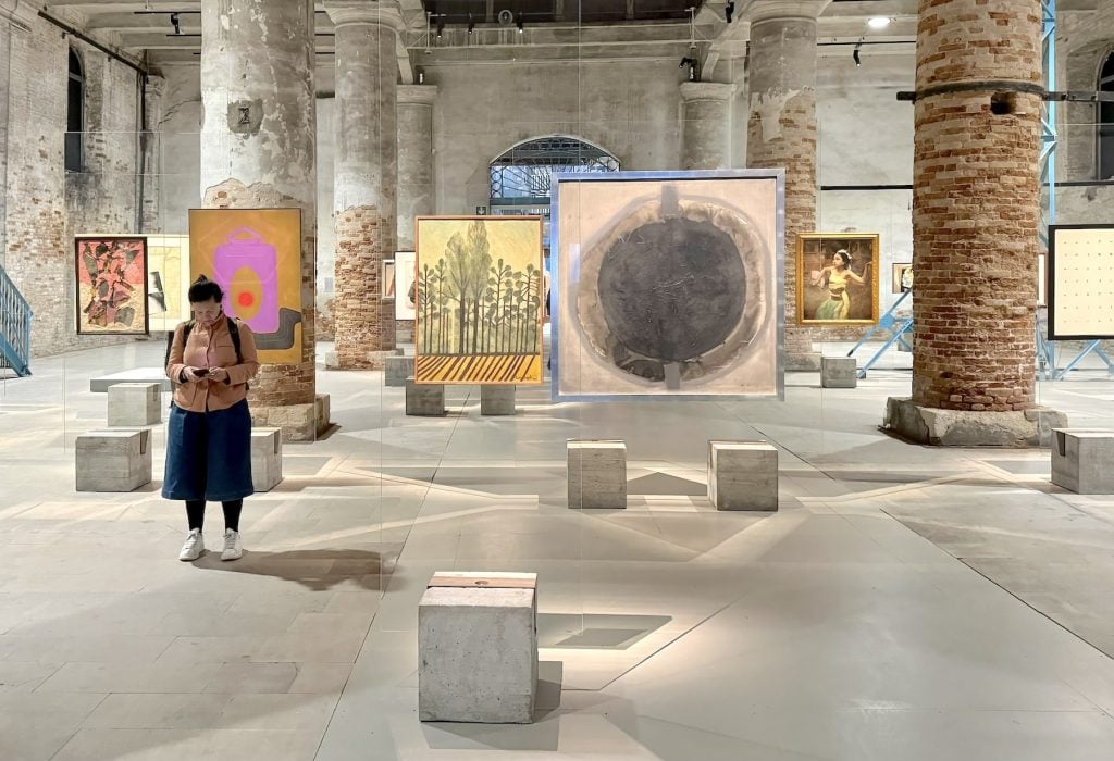 Various paintings are displayed in an art gallery mounted on glass panels connected to concrete blocks for a base