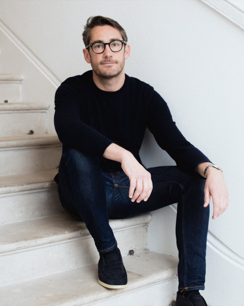 Portrait of Mustard Contemporary director Oliver Munts sitting on white marble steps wearing black long sleeve crew neck shirt and dark wash jeans.
