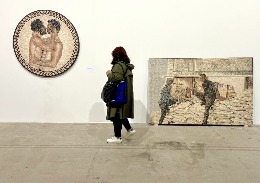 A woman looks at two artworks, made from mosaic tiles, one depicting two men kissing and another depicting two men in front of a museum