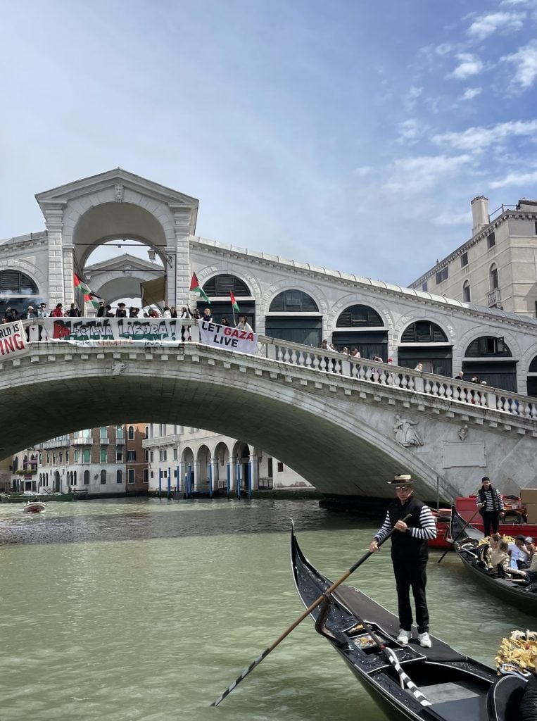 The image is of the Rialto Bridge in Venice. Protestors wave Palestine flags over the bridge. A boat floats underneath.