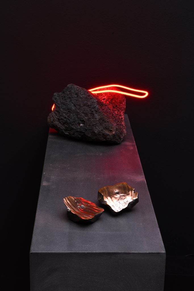 A neon and lava rock sculpture sit on a plinth above two small ceramic sculptures in a darkened room.