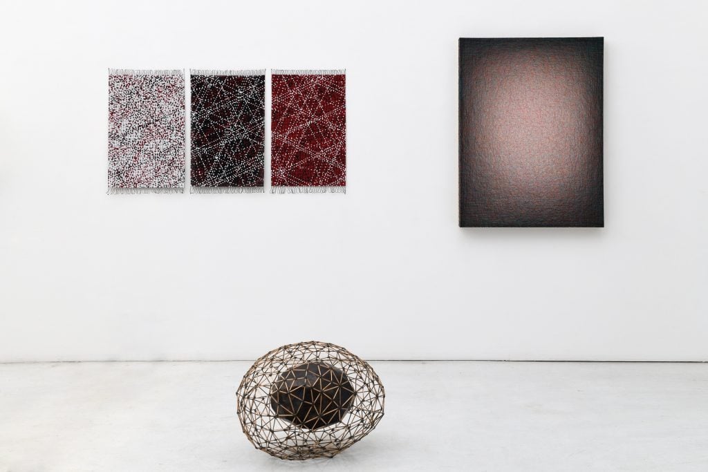 Three mixed media works, two hanging on the wall. The one on the left is comprised of three panels of beadwork in white black and red. On the right, a wire and nails composition on a rectangular frame. And on the ground a wire matrix with a black object suspended in the middle.