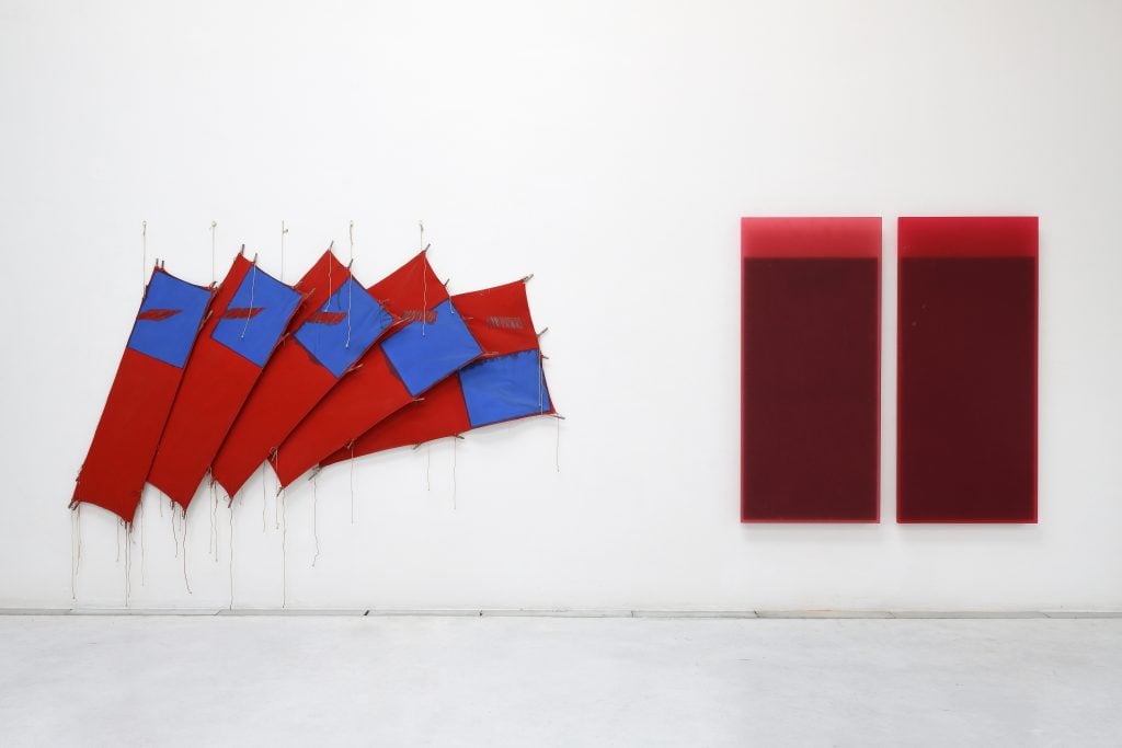 Two wall mounted artworks in a white gallery space, on the left five red fabric panels fan out with swaths of blue. On the right two oxblood panels parallel with bright red strips across their top.