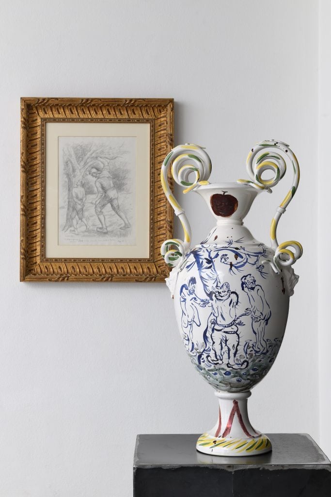 Two artworks, one pencil on paper drawing in the background of a classical vignette, in the foreground a white urn ceramic jug with delicate blue paintings on it.