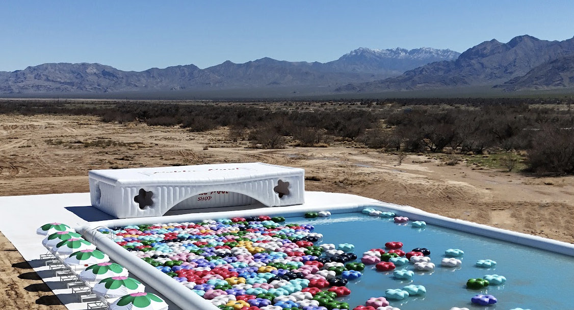 An aerial photograph of a 50 foot long inflatable pool filled with blue water in a desert valley, with multicolored floaties in it and shaded lounge chairs surrounding it