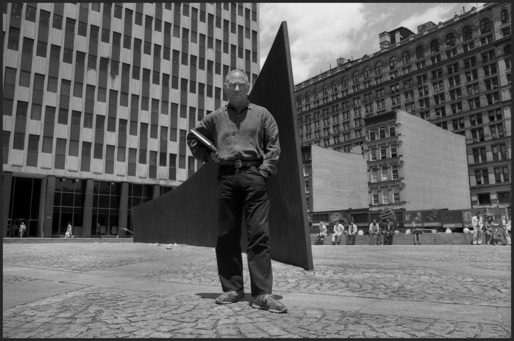 a black-and-white photograph of a white man in a button shirt and pants standing in a outdoor plaza in front of a sculpture that looks like a long steel wall