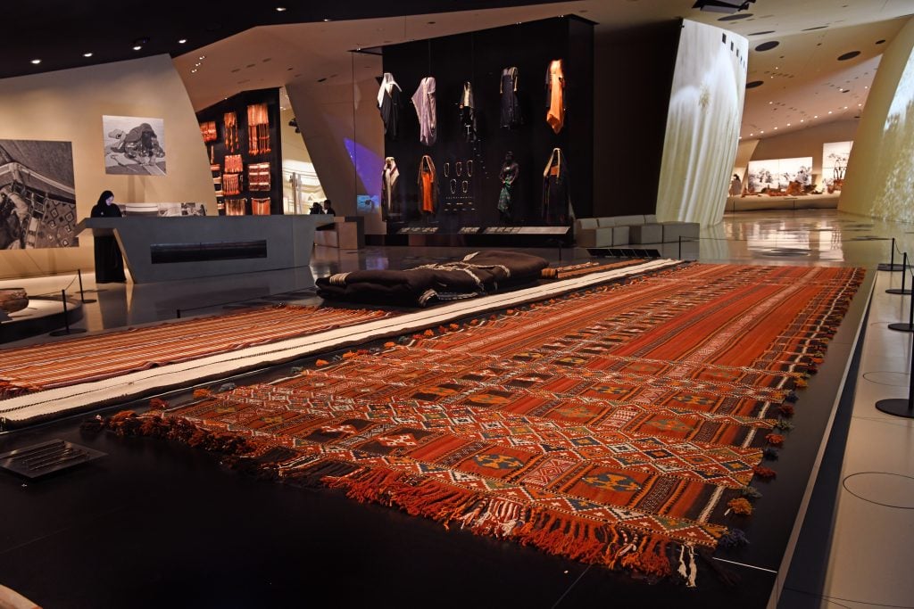 An exhibition gallery featuring a long piece of red Al Sadu textiles and clothing in the background.