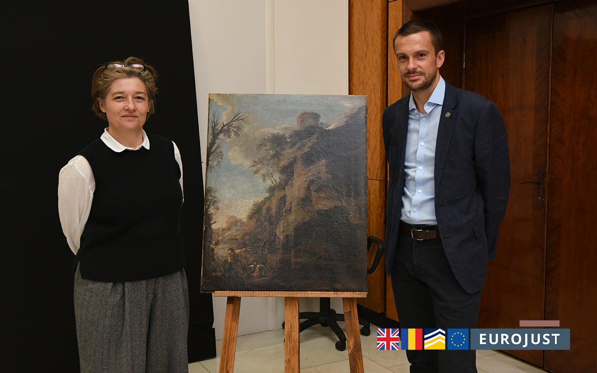 Two individuals are standing beside a painting on an easel, smiling at the camera. The painting appears to be a classic landscape featuring a rocky terrain with figures in a forested area, possibly from the 17th or 18th century. The woman on the left is dressed in a black vest over a white blouse, with her hair tied back and glasses on her head. The man on the right wears a blazer over a button-up shirt and trousers. Behind them is a plain backdrop and a wooden door. The image includes the logo of "Eurojust" with the flags of the UK and EU, suggesting a formal or official context, perhaps related to the recovery or repatriation of the artwork.