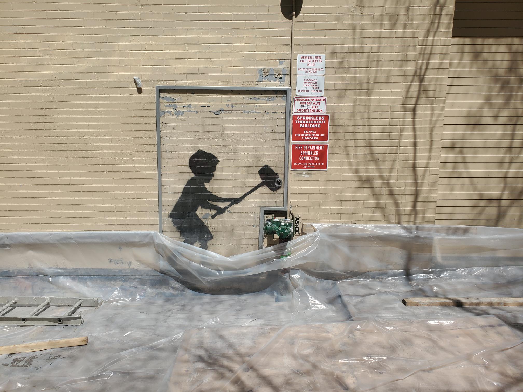 In the image, we see a clear day with a strong shadow casting on a brick wall, indicating bright daylight conditions. A section of the wall is covered with a clear plastic sheet that is wrinkled and partially lifted from the ground, revealing construction or maintenance materials underneath, like wooden planks and a metal frame. Dominating the view is a stencil-style black silhouette of a child wielding a sledgehammer, positioned as if about to strike downwards. This artwork creates a striking contrast with the otherwise nondescript urban setting. Above the plastic sheeting, multiple warning and informational signs are affixed to the wall, providing details about the building's safety and maintenance protocols, such as contact information in case of fire and a notice about the building's sprinkler system. The juxtaposition of the playful, seemingly mischievous artwork with the orderly and serious warning signs creates a thought-provoking visual commentary.