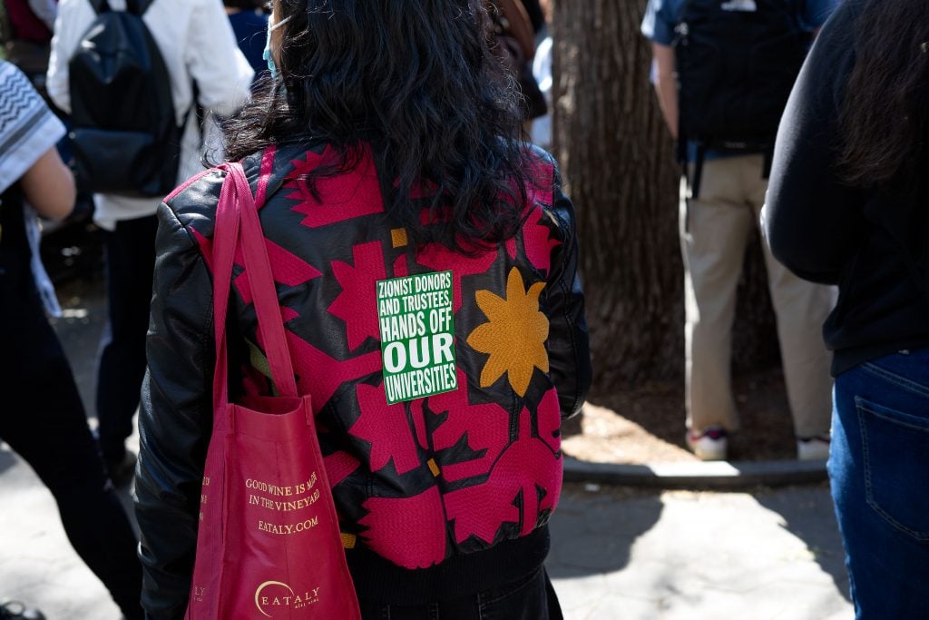 The image shows a person from behind wearing a jacket with a bold design and text that reads, "ZIONIST DONORS AND TRUSTEES, HANDS OFF OUR UNIVERSITIES." The individual is also carrying a red tote bag with a promotional slogan. This scene is set against a backdrop of a bustling crowd, possibly at a public gathering or protest.
