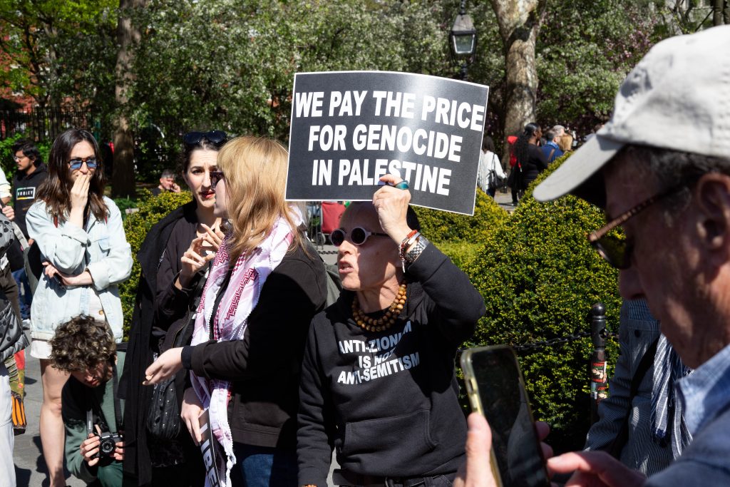 A protestor holds a sign stating "WE PAY THE PRICE FOR GENOCIDE IN PALESTINE" at a rally. In the foreground, other demonstrators engage in conversation, and one wears a sweatshirt with the message "ANTI-ZIONISM IS NOT ANTI-SEMITISM." The scene suggests a public demonstration concerning issues in Palestine, with attendees in casual attire on a sunny day.