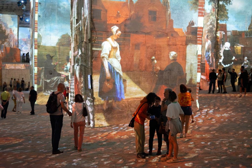 Visitors gather inside a spacious exhibition hall where the walls and floor are covered with large-scale projections of classic paintings, including a prominent image of a woman in period attire, possibly a work by Johannes Vermeer. The ambient lighting casts a soft glow, enhancing the immersive experience.
