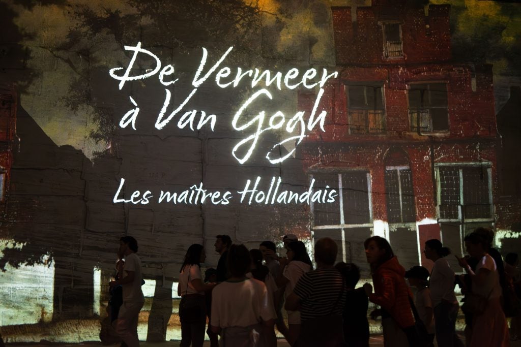 A photo of an immersive art installation, showcasing a wall illuminated with a projected image of a text that reads "De Vermeer à Van Gogh, Les maîtres Hollandais" in stylized white script, indicating a theme focused on Dutch masters Vermeer and Van Gogh. The backdrop is a large, projected image of a traditional Dutch building, with visible architectural details and a tree's silhouette. A group of attendees is seen engaging with the exhibit, with some in silhouette against the bright projection, providing a sense of scale and the communal experience of the exhibition.