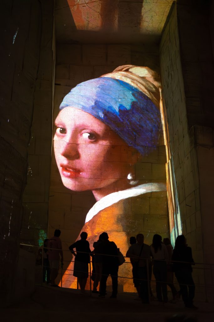Spectators are silhouetted against a towering projection of Johannes Vermeer's "Girl with a Pearl Earring" on a wall within an immersive exhibit space. The artwork's vivid colors and the subject's enigmatic gaze are dramatically enlarged, enhancing the painting's impact.