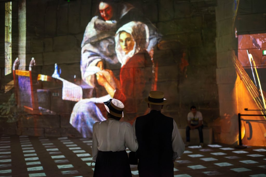 Two visitors dressed in period costumes, reminiscent of the attire seen in classic paintings, stand before a large wall projection of a historic painting, depicting figures in a domestic setting. The projection casts a soft, colorful light across the checkerboard floor of the exhibit space.