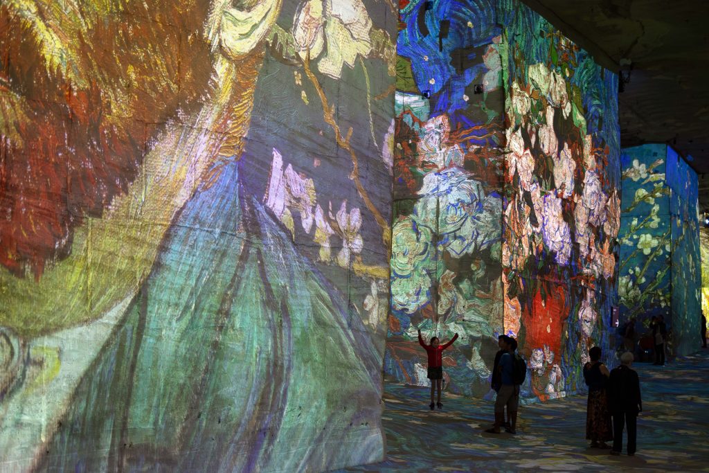 In a vibrant immersive art installation, visitors are surrounded by towering walls filled with large-scale projections of Van Gogh's expressive paintings, featuring his iconic swirls and bold brushstrokes in a multitude of colors. One person stands with arms outstretched towards the artwork, while others observe the captivating scenes.