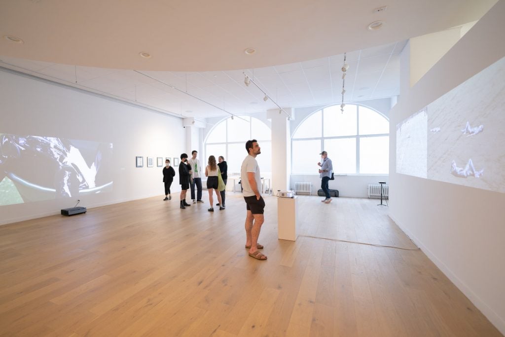 Inside a white room gallery space with a large window at the end and people milling about inside, the site of exhibitions for Sotheby's Institute of Art.
