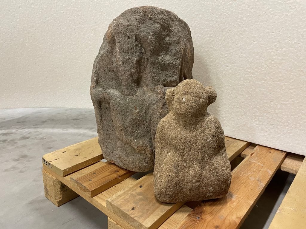A photograph of two stone sculptures sitting atop a wooden pallet in a beige room