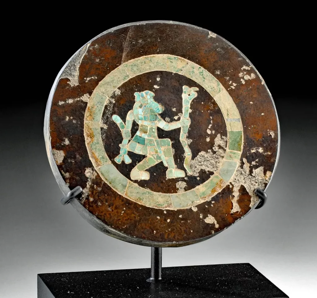 A Pre-Columbian disc with a mosaic of what seems to be turquoise forming the figure of a person or deity holding a staff, surrounded by a circular motif. This type of work is reminiscent of ancient Mexican cultures, potentially from the Mixtec or Aztec peoples.