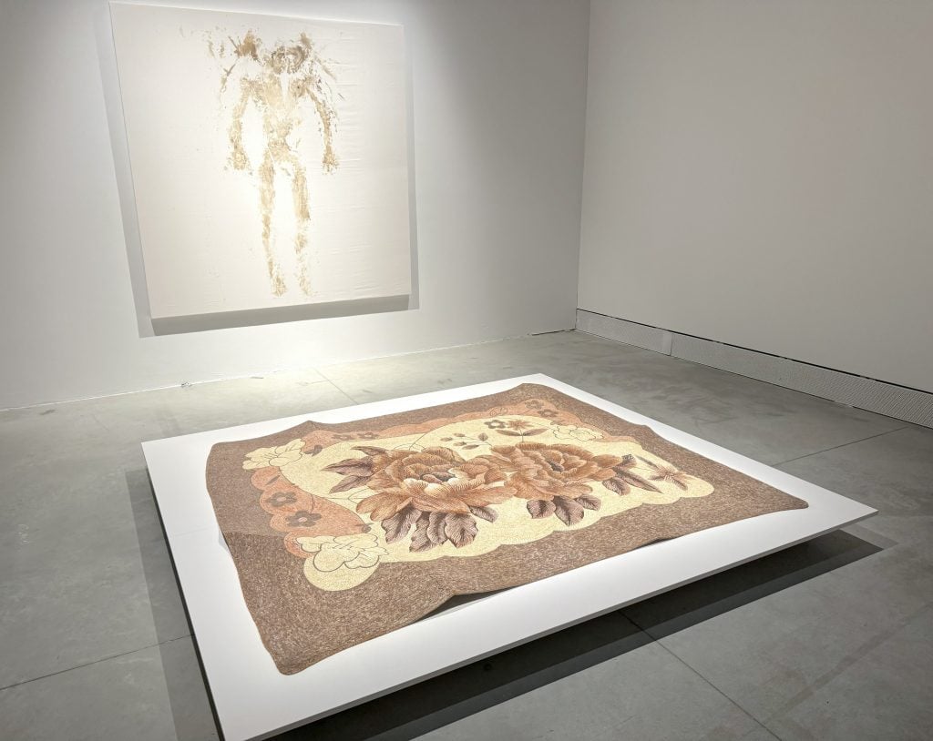 An image bearing the imprint of a human body hung on the wall in front of a blanket recreated in mosaic tiles
