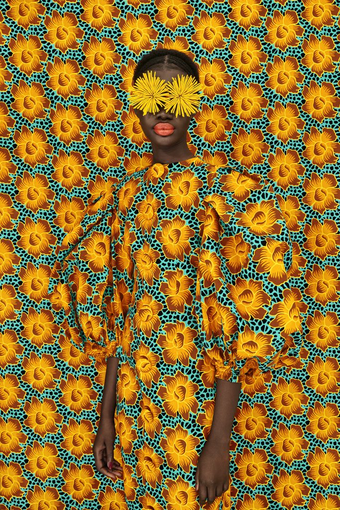 A figure in a bright patterned dress that blends in with the background that is the exact same bright floral pattern, with two spider mums covering their eyes.