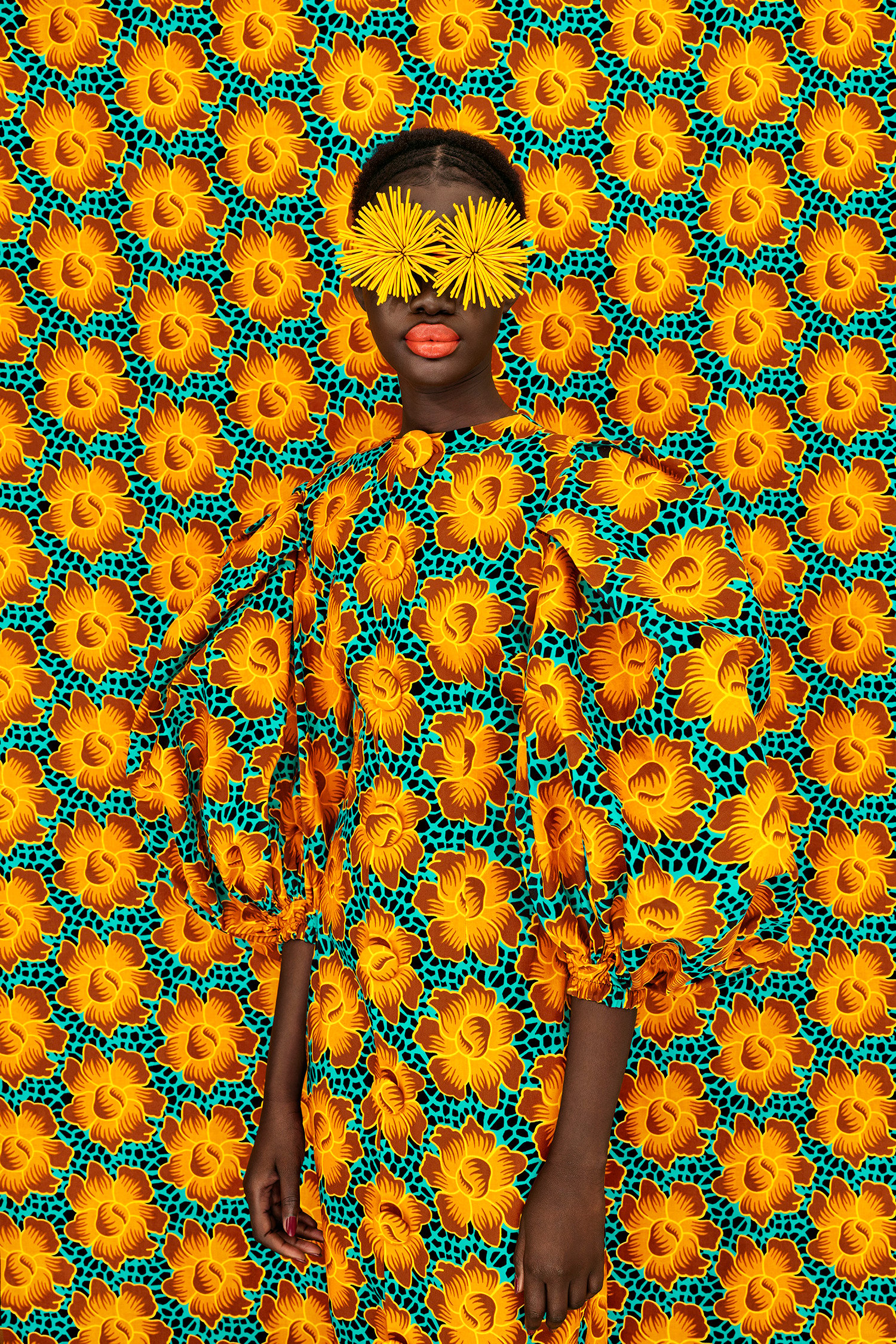 A figure in a bright patterned dress that blends in with the background that is the exact same bright floral pattern, with two spider mums covering their eyes.