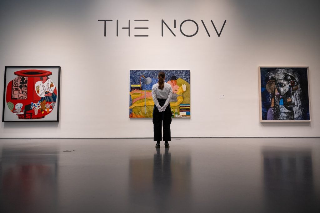 a person stands with their back toward the viewer in front of contemporary paintings hung on a white wall