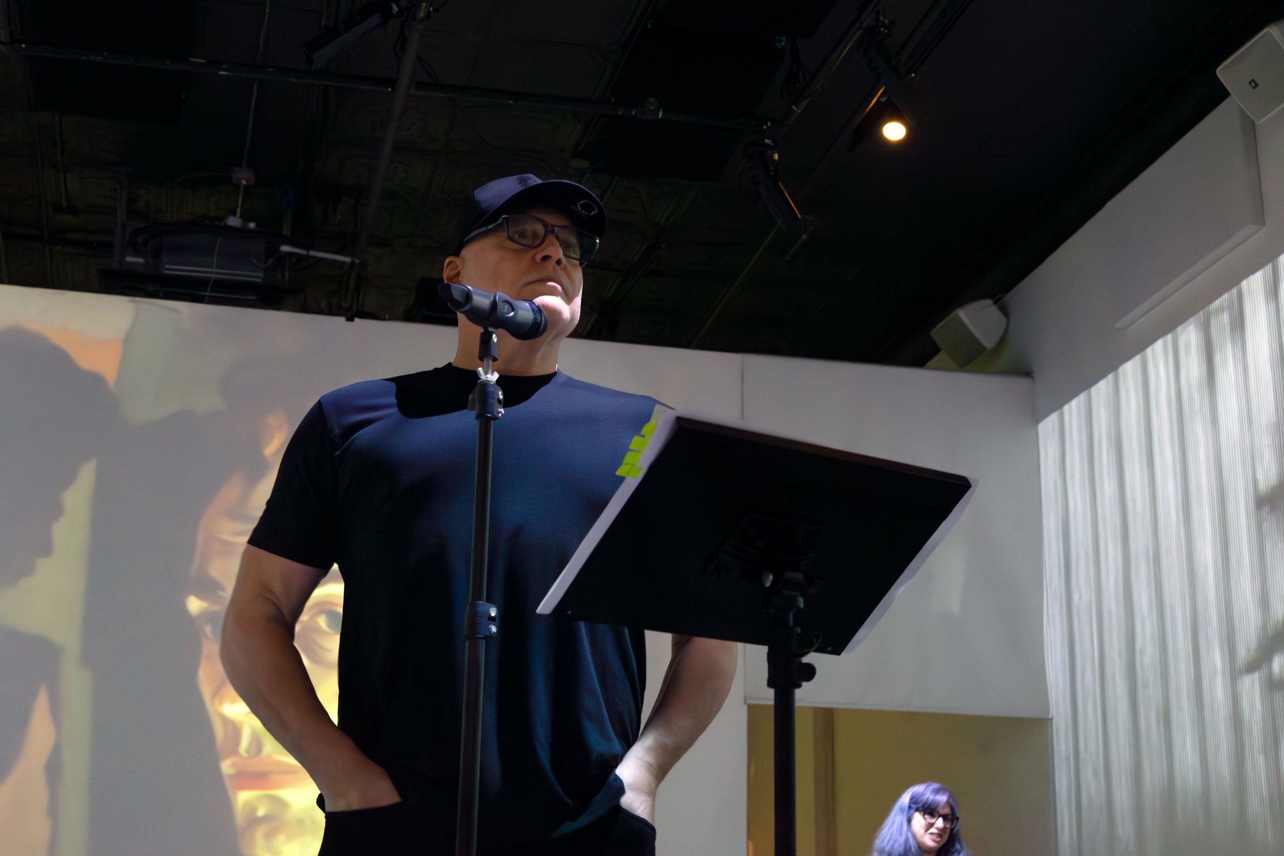 A man, actor Vincent D'Onofrio, stands at a microphone reading from a music stand with pages that appear to have notes or text