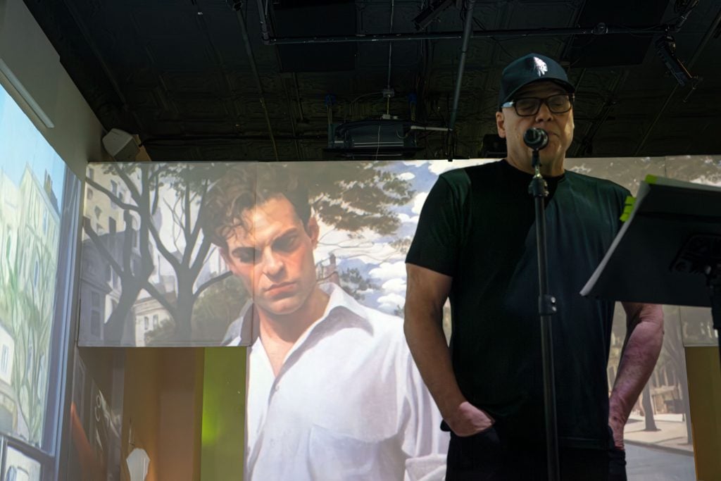 A man, actor Vincent D'Onofrio, in a dark shirt and cap standing at a microphone in front of a large projected image of a painting of another man.