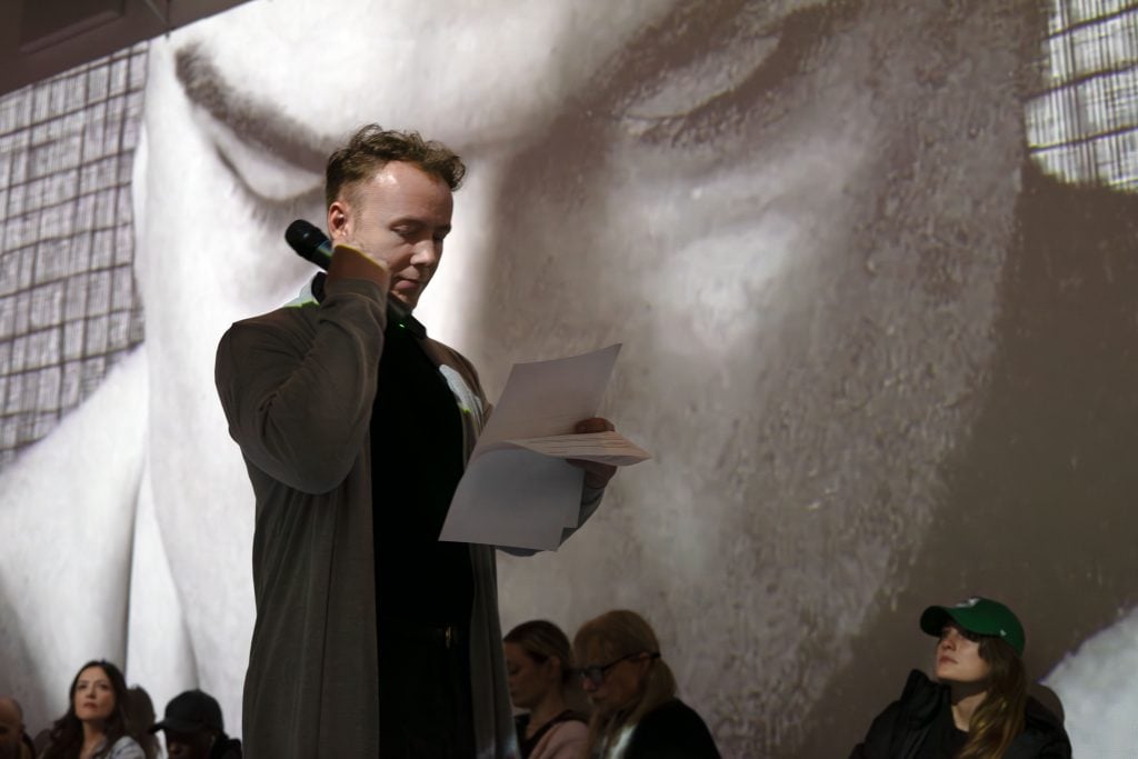 A man, dressed in a formal long coat and holding a microphone close to his chin, appears to be reading from papers in his hand, perhaps delivering a speech or a reading. His attention is focused on the sheets, suggesting concentration on the material. The large projected background showcases an extreme close-up of what seems to be an artwork or photograph, emphasizing the texture and detail of an eye. This striking visual element adds a dramatic backdrop to the speaker's activity. Audience members are visible in the lower frame, looking up towards the speaker or the projection, indicating engagement with the performance. The setting appears to be an indoor event where art and spoken word are central to the experience.